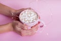 Female hands with French manicure holding a pink cup of cacao with marshmellow. Pink background with copy space