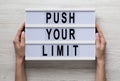 Female hands hold a lightbox with `Push your limit` words over white wooden background, top view. Overhead, from above, flat lay