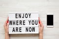 Female hands hold modern board with `Enjoy where you are now` word, smartphone with blank screen on white wooden background, top