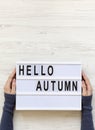 Female hands hold modern board with text `Hello Autumn` over white wooden surface, top view. Overhead, flat lay