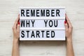 Female hands hold modern board with text `Remember why you started` over white wooden background. From above, flat-lay