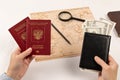 Female hands hold two passports and a black leather wallet with money. on a white wooden table lies a map of the world, a magnifyi