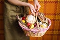 Female hands holding basket with colorful eggs, cake, red wine, hamon or jerky and dry smoked sausage. Food gift set for