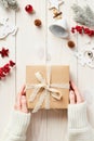 Female hands holding craft paper gift box with ribbon bow over white wooden plank desk table decorated Christmas ornaments. Royalty Free Stock Photo