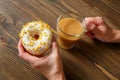 Female hands holding cup of coffee and donut in a white glaze with yellow confectionery sprinkles on a wooden background Royalty Free Stock Photo