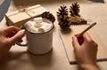 Female hands holding a cup of hot cocoa drink and marshmallows and writing on blank paper sheet of notebook with a wooden pencil Royalty Free Stock Photo