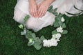 Female hands holding a decorated wreath of flowers and leaves on a background of green grass. Top view. Copy, empty