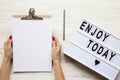 Female hands holding an empty noticepad over white wooden background, top view. `Enjoy today` word on lightbox. Royalty Free Stock Photo