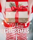 Female hands holding gift box with red ribbon and Merry Christmas and New Year typographical on shiny xmas background.