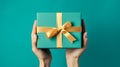 Female hands holding gift box tied with gold ribbon teal background. Holiday presents shopping celebration