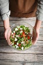 Female hands holding a glass bowl of vegetable salad with feta over a wooden background