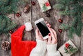 Female hands holding mobile phone on wooden background with Christmas gifts, Santa hat, Fir branches