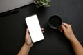 A female hands holding a smartphone mockup and a black coffee cup over modern black tabletop
