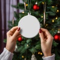 female hands holding white blank christmas ball on tree background, AI Generated Royalty Free Stock Photo