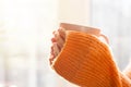 Female hands with hot drink. A young woman in orange sweater holding cup of tea or coffee near window at home Royalty Free Stock Photo