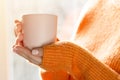 Female hands with hot drink. A young woman in orange sweater holding cup of tea or coffee near window at home Royalty Free Stock Photo