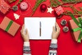 Female hands in knitted sweater are writing with pen in clean notebook plans for the new year, gift boxes, fir branches on red bac Royalty Free Stock Photo