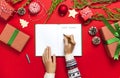 Female hands in knitted sweater are writing with pen in clean notebook plans for the new year, gift boxes, fir branches on red bac Royalty Free Stock Photo