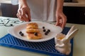 Female hands putting blueberry on traditional syrniki or cottage cheese pankaces and honey pot with blue napkin on kitchen table.