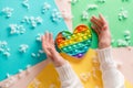 Female hands surround a silicone rainbow antistress toy in the shape of a heart on a cardboard of different colors and fluff.