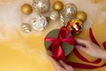 Female hands tie a red ribbon on a green round gift. Christmas background with golden balls and angel hair. Top view Royalty Free Stock Photo