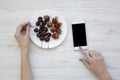 Female hands using smartphone while eating cherries and strawberries on white wooden background, top view. Royalty Free Stock Photo