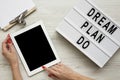 Female hands using tablet with blank screen, modern board with text `Dream plan do`, clipboard over white wooden background, Royalty Free Stock Photo