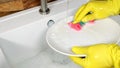 Female hands wearing yellow rubber gloves washing white plate in kitchen sink with foam sponge. Royalty Free Stock Photo