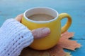 Female hands in white sweater holding yellow cup of coffee on blue wooden table with autumn leaves background. Royalty Free Stock Photo