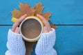 Female hands in white sweater holding yellow cup of coffee on blue wooden table with autumn leaves background. Royalty Free Stock Photo