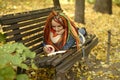 Female hipster writing in diary on bench in park. Young dreamy woman with long dreadlocks lying on wooden bench and Royalty Free Stock Photo