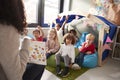 Female infant school teacher sitting on a chair showing a book to a group of children sitting on bean bags in a comfortable corner