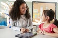 Female infant school teacher working one on one with a Chinese schoolgirl, sitting at a table in a classroom using a tablet comput