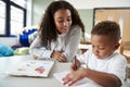 Female infant school teacher working one on one with a young schoolboy, sitting at a table writing in a classroom, front view, clo Royalty Free Stock Photo