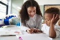 Female infant school teacher working one on one with a young schoolboy, sitting at a table writing with him, close up