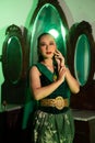 a female Javanese dancer in a green dance costume and a full face with makeup poses with her hands in front of a dressing table