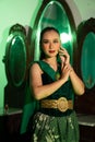 a female Javanese dancer in a green dance costume and a full face with makeup poses with her hands in front of a dressing table