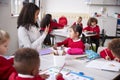 Female kindergarten teacher sitting at table in a classroom talking and gesturing to a young Chinese schoolgirl, selective focus