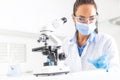 Female lab technician in protective glasses, gloves and face mask sits next to a microscope and conical flask, looking aside on Royalty Free Stock Photo