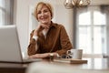 Female With Laptop. Senior Blonde Woman In Brown Jacket Sits At The Desk With Laptop And Cup.