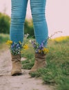 Female legs in jeans high boots with inserted flowers on the road in the field Royalty Free Stock Photo