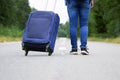 Female legs in jeans and sneakers with a travel bag on wheels walks away on the road Royalty Free Stock Photo