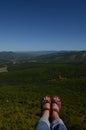 Female legs in trekking shoes over a huge precipice against a background of green forest and blue sky. Crimea