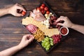 Female and male hands taking snacks from charcuterie board with sausage, fruit, crackers and cheese on a dark wooden background Royalty Free Stock Photo