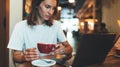 Female manager holding in hands cup of coffee using laptop in cafe, girl freelancer looks at monitor while relaxing Royalty Free Stock Photo