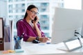Female manager talking on cellphone and taking notes, sitting at her desk in office, working at table with computer Royalty Free Stock Photo