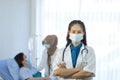 Female medical doctor wear face mask arms crossed stand in hospital with medical team working on the background Royalty Free Stock Photo