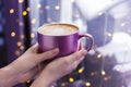 Female nands holding a lilac mug of coffee on a dark background with golden bokeh.