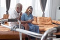 Female nurse takes care of senior patient and teaches him to use digital tablet at living room house, elderly mature grandfather Royalty Free Stock Photo