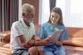 Female nurse takes care of senior patient and teaches him to use digital tablet at living room house, elderly mature grandfather Royalty Free Stock Photo